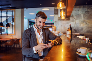 Happy businessman in elegant blazer sitting in the modern concept bar and using his phone to text a message or make a phone call. Telecommunications, social media, stay connected