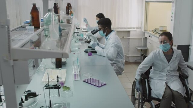 Slowmo side view shot of disabled female scientist in wheelchair wearing white coat, gloves and face mask and putting solution into microplate while her colleagues working on research in lab