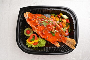 deep fried crispy whole grouper fish seafood with sweet and sour chilli sauce and lychee fruits on top in black bento tray box for asian take away halal menu
