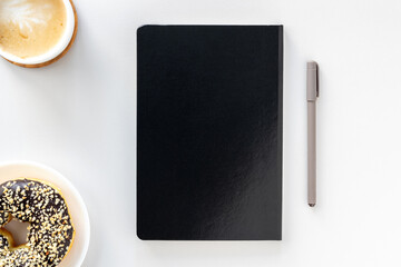 Flat lay of workspace desk, black notepad, pen, coffee and chocolate donut