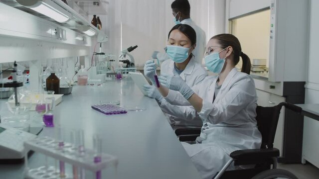 Side view slowmo shot of disabled female chemist in wheelchair and her female colleague discussing solution in test tube while working in lab Male black scientist working in background