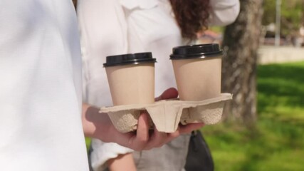 Mid-section tracking shot of unrecognizable man holding disposable takeaway coffee cups tray and walking beside woman on sunny summer day