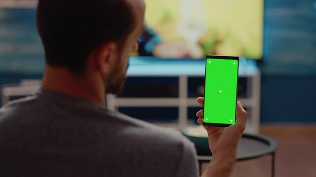 Person holding mobile phone with green screen vertically on display sitting in living room at home. Isolated background with chroma key and mockup for media template on copy space