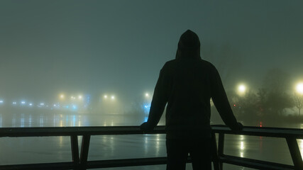 A hooded figure silhouetted against street lights next to a river. On a mysterious misty, winters...