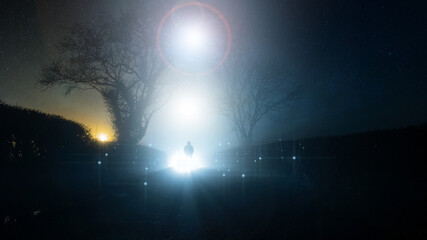 A supernatural, UFO concept of a scary, mysterious hooded figure, standing in front of a bright lights in the sky, on a country lane, on a spooky foggy winters night.