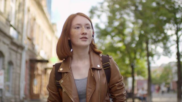 Slowmo tracking shot of ginger teenage girl in earphones holding mobile phone and walking down street while talking to someone or singing