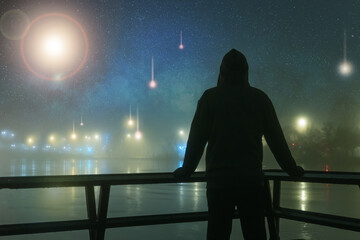 A hooded figure silhouetted against alien, UFO, spaceship lights next to a river. On a mysterious misty, winters night, with a universe of stars. Science fiction concept.