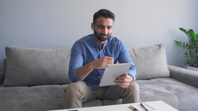 Millennial indian man using digital tablet on sofa and smiling slow motion
