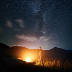 Side view of man standing and enjoying near fire and relaxing under starry sky. Concept of...