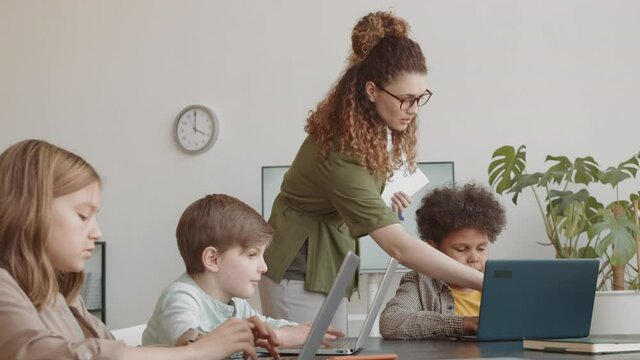 Tracking left medium long of young Caucasian woman wearing glasses, teaching diverse elementary-aged boys and girl sitting at table, using computers on lesson