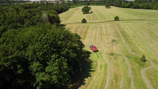 Aerial drone tracking shot of tractors pulling trailers and a forage harvester collecting grass cuttings for silage from the edge of a tree lined field
