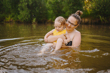 Fototapeta premium Little funny cute blonde girl child toddler in yellow bodysuit laughing learns swim outside at summer lake. Plus size body positive smiling woman mother baby swimming in natural pool. Water splashes