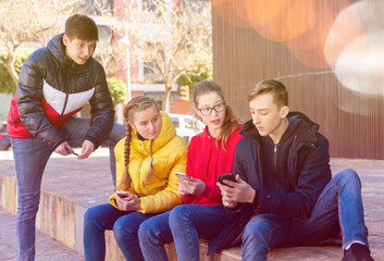 Four teenagers having friendly discussion and using cellphones during gathering outdoors on spring day