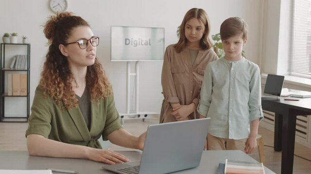 Medium long of two guilty Caucasian boy and girl standing by desk of young angry Caucasian teacher, lecturing students for bad homework