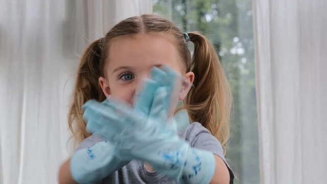 Concentrated little preschooler girl with long pigtails puts on large sterile gloves and claps hands at home close view