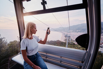 Traveling by Turkey. Young tourist woman sitting in Alanya's cable car enjoying view taking photo on her smartphone.