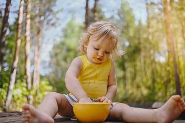 Little funny cute blonde girl child toddler with dirty clothes and face eating baby food fruit or vegetable puree with spoon from yellow plate outside at summer. Healthy happy childhood concept