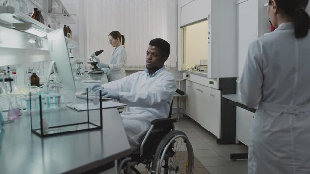 Slowmo tracking of disabled black male scientist in wheelchair working on computer in lab, then posing His female colleagues doing their research in background White lab rat is in glass tank