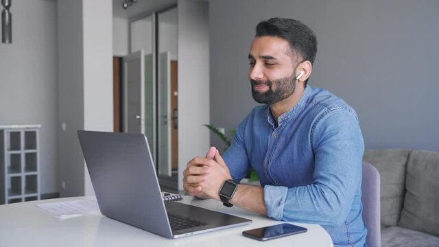 Adult indian man wearing wireless earphones make video call looking at laptop