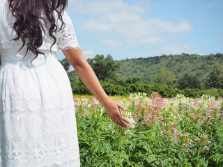 Woman white dress in flower park and touching flower near her.Asian woman traveler with pink flower park and mountain and blue sky background.