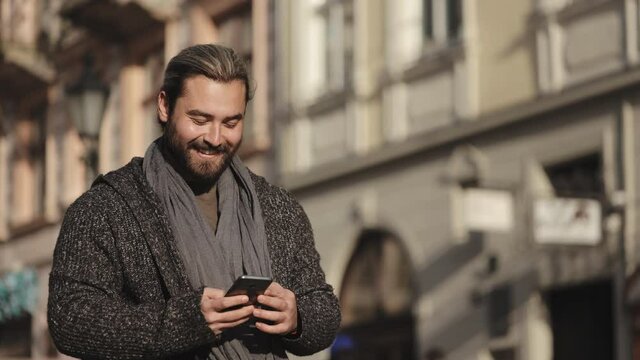 A happy bearded man is walking and texting on his smartphone. He is smiling and looking around. He's wearing a sweater and scarf. The sun is shining. 4K