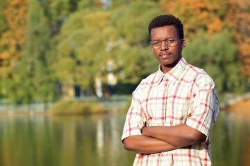 Portrait of serious handsome black African Afro American ethnic guy, young man in glasses standing in golden autumn, fall park with his hands, arms crossed, looking at camera in shirt near lake