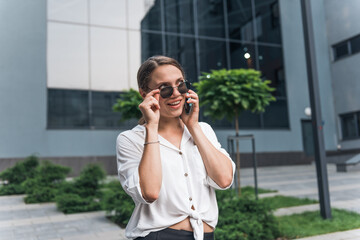 Business woman wearing trendy sunglasses walks down the central city street and uses her phone. 