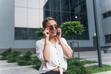 Fototapeta premium Business woman wearing trendy sunglasses walks down the central city street and uses her phone. 