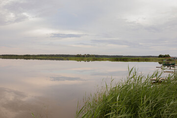 The mirror-like surface of the lake on the outskirts of the village with old piers and boats. Cloudy weather, evening.