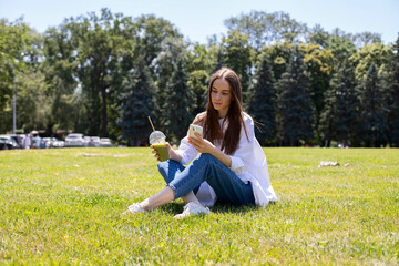 A young girl is sitting in the park on the grass and drinking a green smoothie