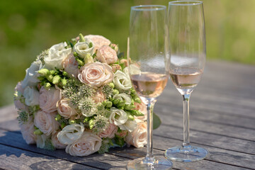 Two romantic elegant flutes of pink champagne alongside a bridal bouquet with matching pink and white roses on a rustic wooden table outdoors at a wedding reception in a close up low angle view