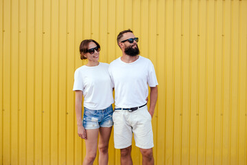 Young smiling pretty happy cheerful positive couple in blank white t-shirts near color background on street. Mockup for design.