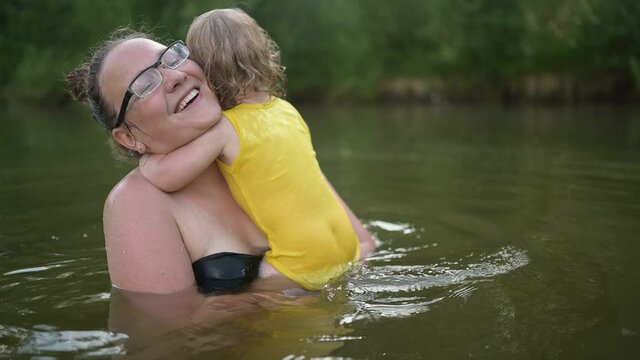 Little funny cute blonde girl child toddler in yellow bodysuit laughing learns swim outside at summer lake. Plus size body positive smiling woman mother baby swimming in natural pool. Water splashes