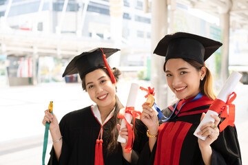 Two Asian women who wore graduate gowns are looking forward to the camera and smiling widely with certificates in their hands while another hand holding medals.