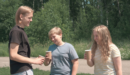 caucasian teenagers having fun drinking coffee to go in a park. talking and laughing boys and girl, one very tall boy. friends on vacation outdoors, summer sunny day.