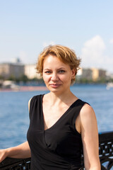 Fototapeta premium Portrait of a young girl with a short haircut. A girl on a sunny day poses on the embankment in a black dress.