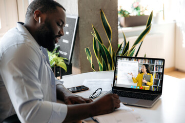 Online education. Focused african american guy sits at the table at home, listens to an online...
