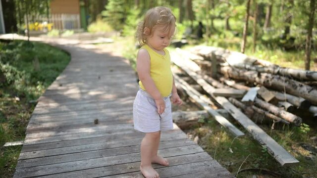 Little funny cute blonde girl child toddler in yellow undershirt and grey shorts walking playing on wooden eco trail outside at summer at front yard countryside. Healthy happy childhood concept. 