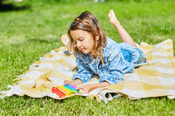 Girl lies on a blanket on grass outdoors and play pop it, kid hands playing with colorful pop It, fidget toy in the backyard of the house on a sunny summer day, summer time vacation.