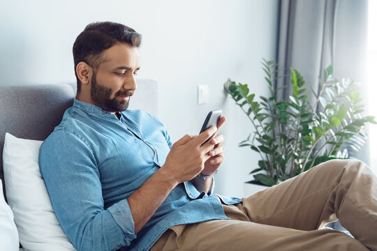 Handsome young adult indian man using smartphone while lying on sofa at home