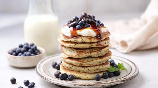 Pouring maple syrup on stack of healthy poppy seed pancakes served with greek yogurt and wild blueberries