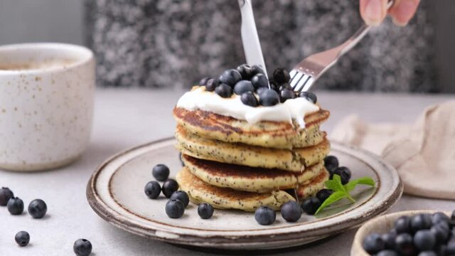 Young woman eating poppy seed pancakes with yogurt and blueberries using fork and knife, cutting stack in halves