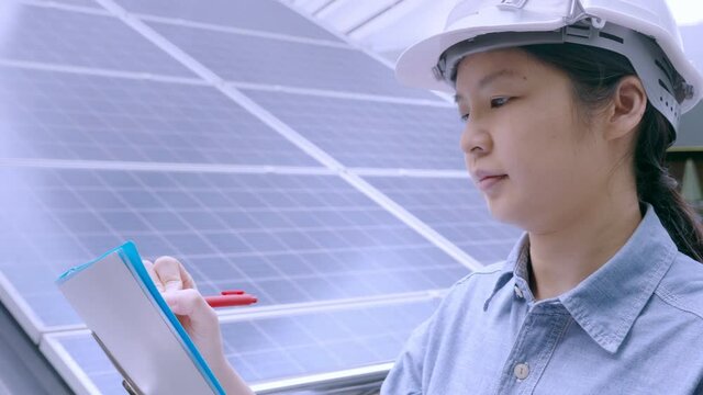 Side view, close up of Asian female engineer wearing safety hard hat, working outdoors near solar rooftop panel, holding clipboard and a pen, writing. Solar energy and sustainable business concept