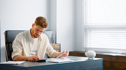 Thoughtful guy manager with beard sums up administrative expenses on calculator