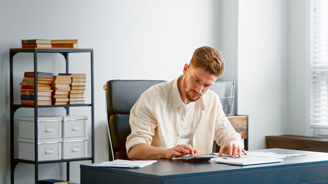 Professional accountant with beard counts paper checks summarizing sums on calculator with serious face sitting at table