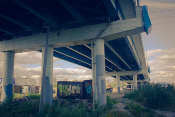 the space under the bridge, a neglected place with graffiti and debris, concrete columns support...