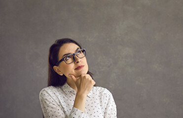 Thoughtful young caucasian woman touching chin by hand studio headshot. Pensive smart girl wearing eyeglasses thinking over good idea or planning something standing and looking up. People emotion