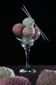 Balls Of Multi-colored Yarn And Knitting Needles Lie In A Large Glass Of Margarita. Hobby Concept. Handmade. Vertical Photo.Black Background.