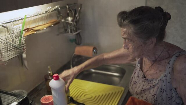 Lonely sad senior woman 90 years old with gray hair forced to wash dishes with her hands due to poverty at home in an old kitchen. Grandmother at work. Old dirty house, poor living conditions.