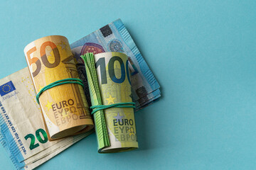 Green roll of 100 euro banknotes and orange roll of 50 euro bills over 20 euro paper money against blue background. Counting money, income and expenses and cash savings concepts. Copy space.